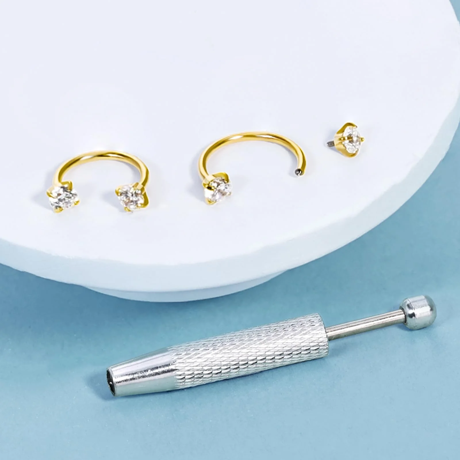Two gold horseshoe rings with clear gems and a small stud on a white disc, with the steel opening tool laid beside them on pale blue