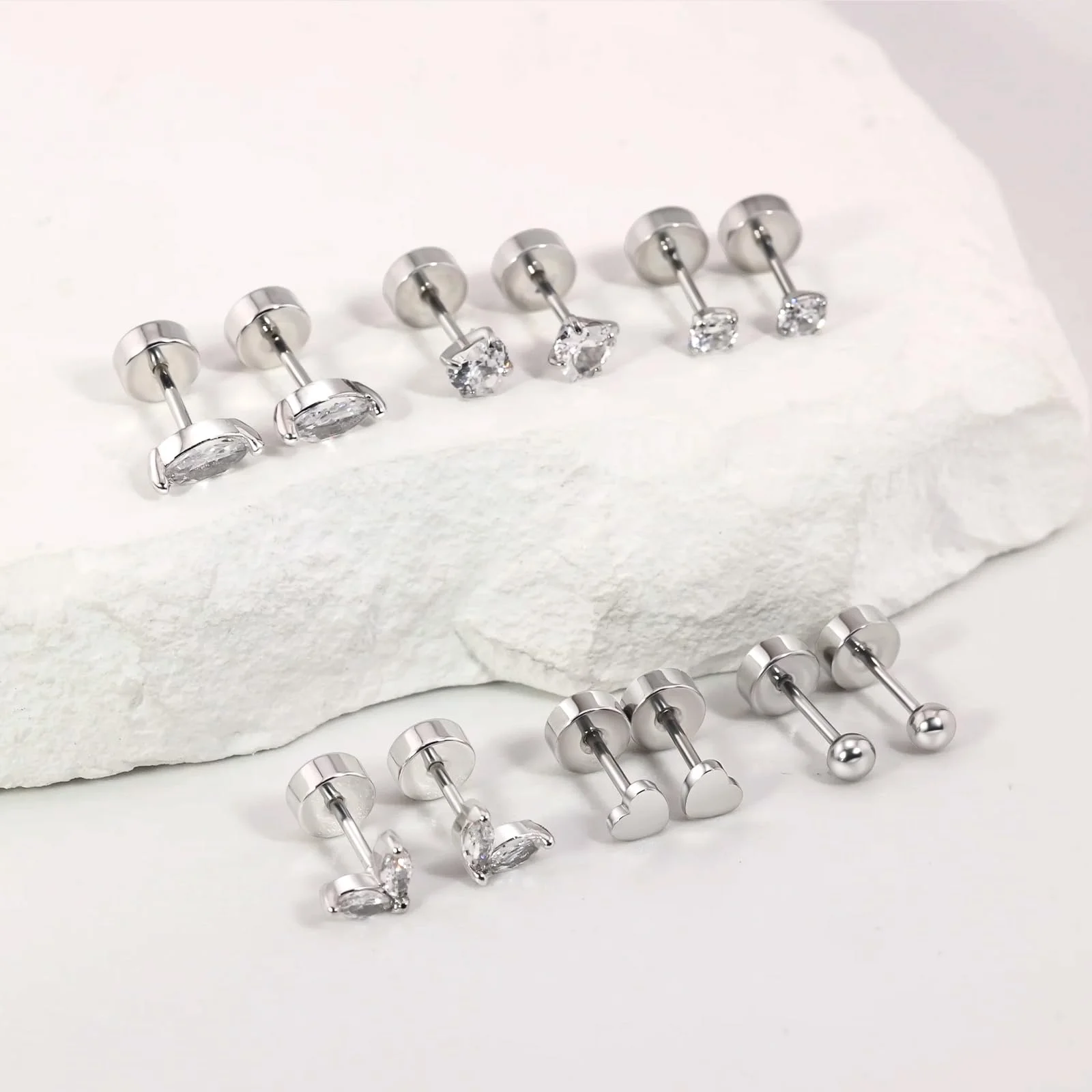 Eight silver flat back studs standing on and beside a piece of white stone, teardrop, round, heart and ball fronts with their discs showing