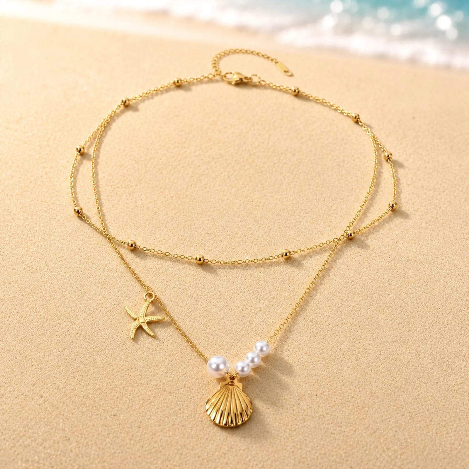 A gold layered chain with a starfish, a row of pearls and a shell charm, laid out on wet sand at the edge of the water
