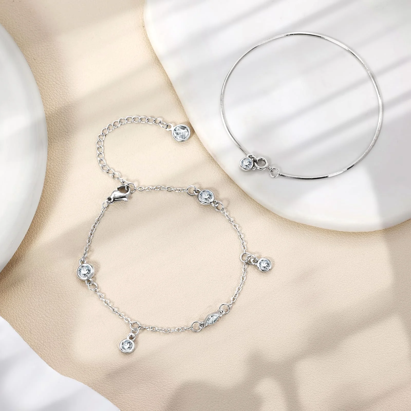 A silver bracelet set with clear round stones next to a plain silver bangle on cream cloth, both throwing a soft shadow