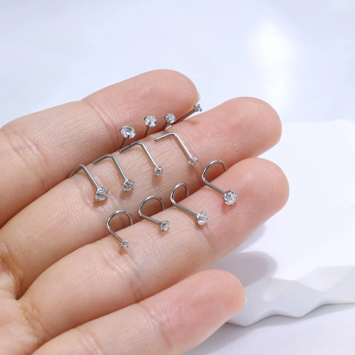 Six silver L-shaped nose studs balanced across four fingers, the bends of the posts standing out against the skin