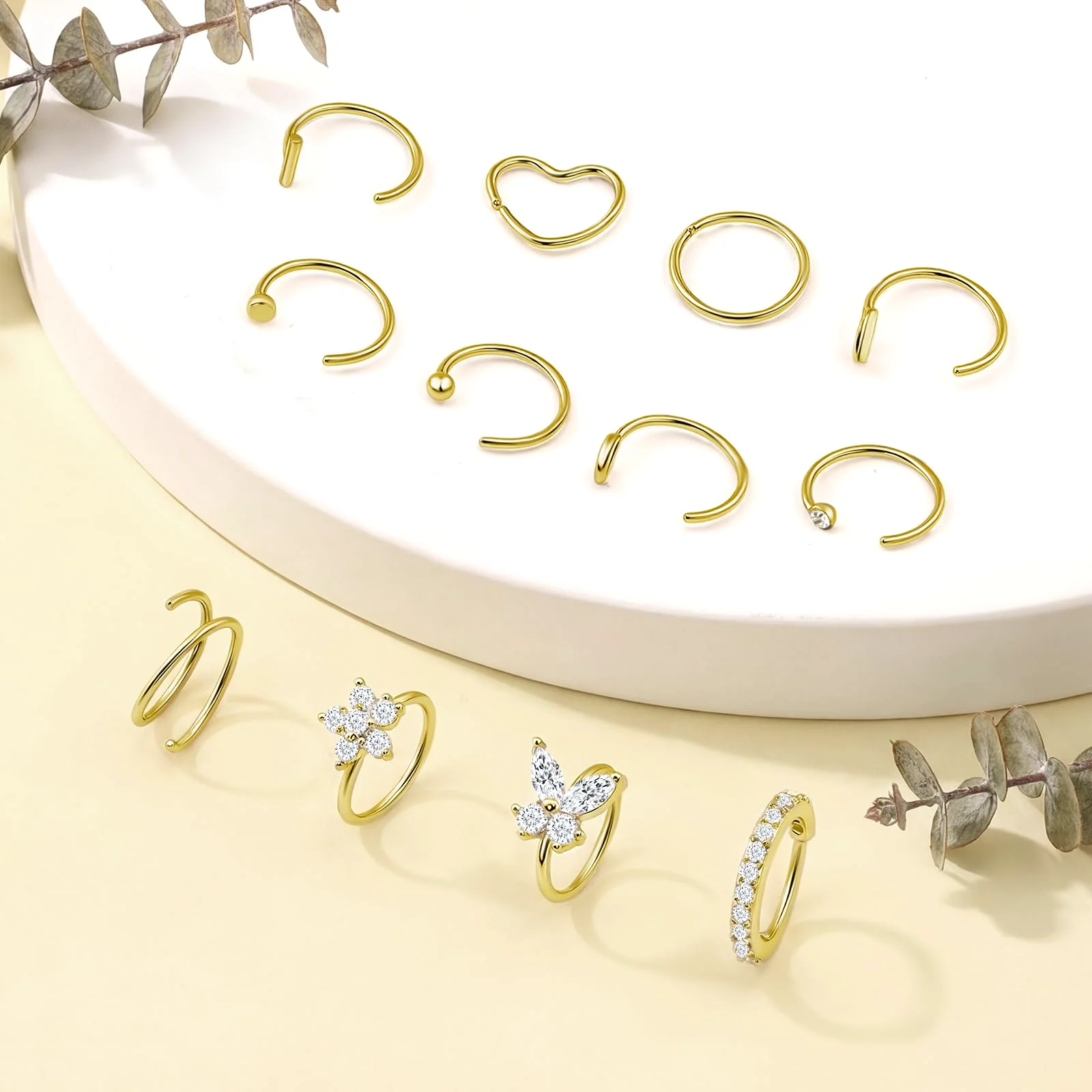 Nine gold nose hoops on a white dish, one bent to a heart, with three paved rings resting on the yellow ground below