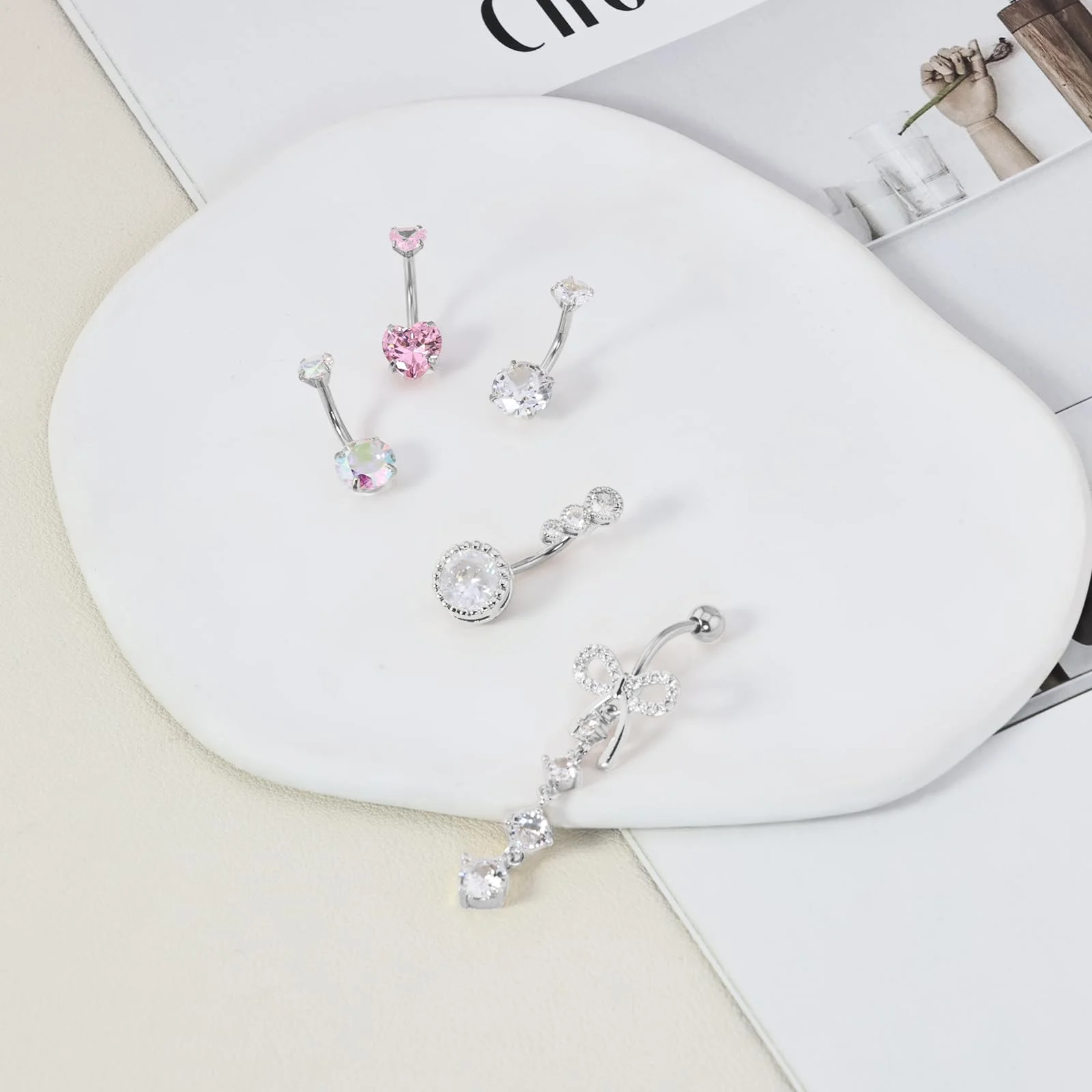 Four silver navel bars on a white dish, one with a pink stone, one with a flower drop and two with clear round gems