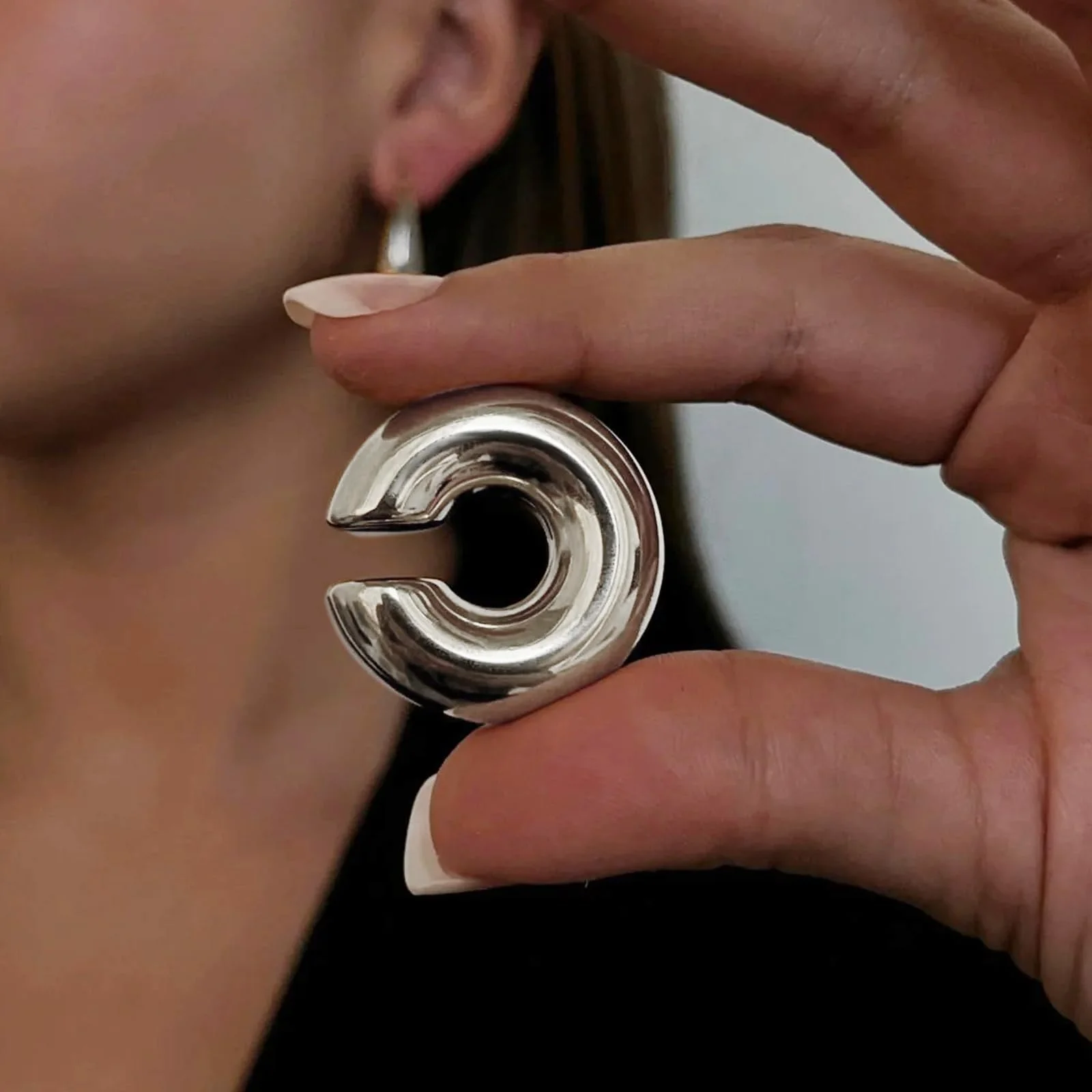 A hand holding a heavy silver ear cuff up beside an ear, the curl of the metal filling the frame