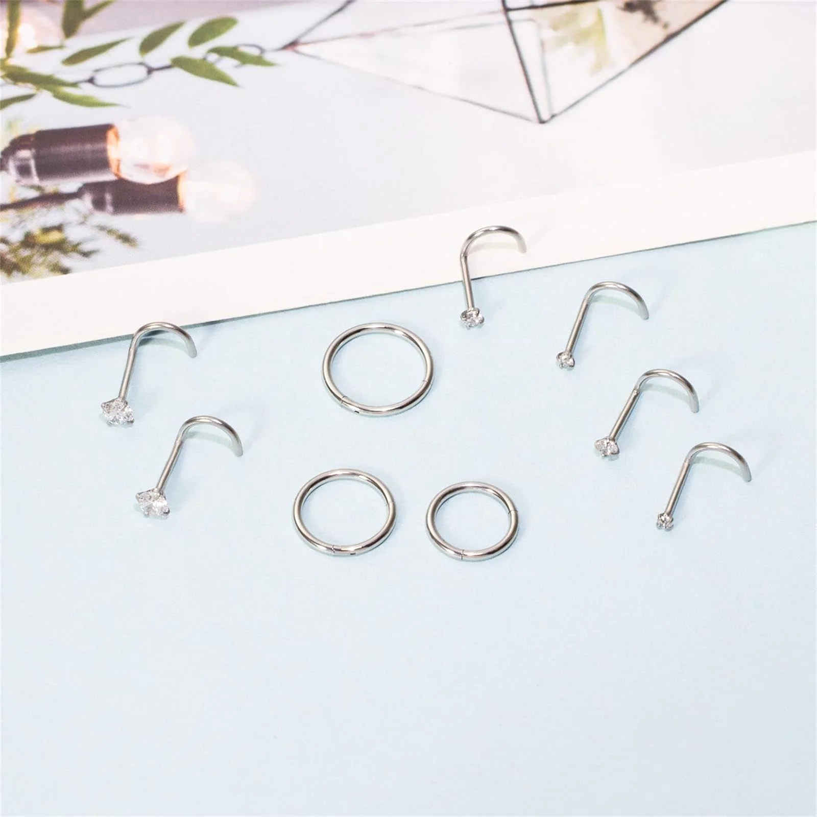 Nine silver pieces laid on pale blue paper: four curved nose studs, one straight post and three plain hoops, with a print propped behind them