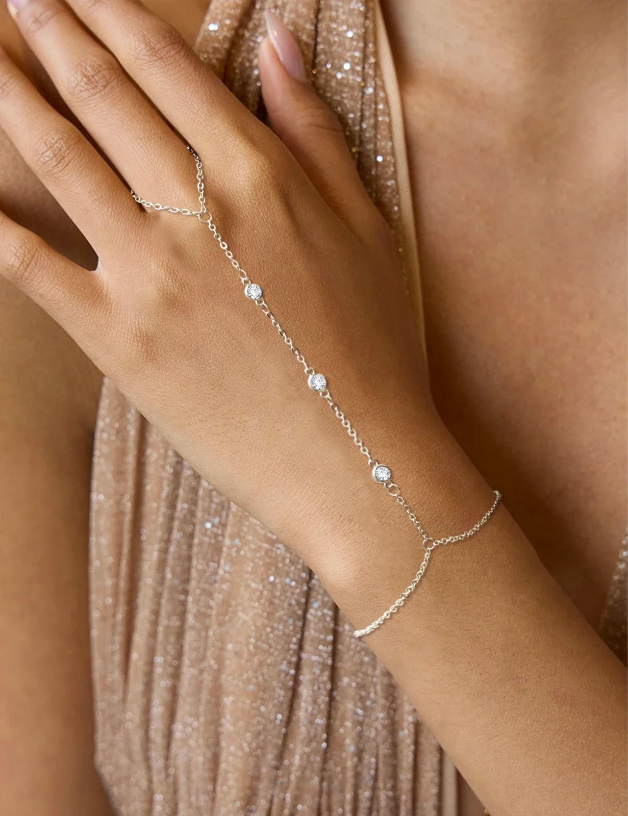 A silver hand chain worn on a hand held against a champagne dress, the chain running from the wrist to a ring on one finger